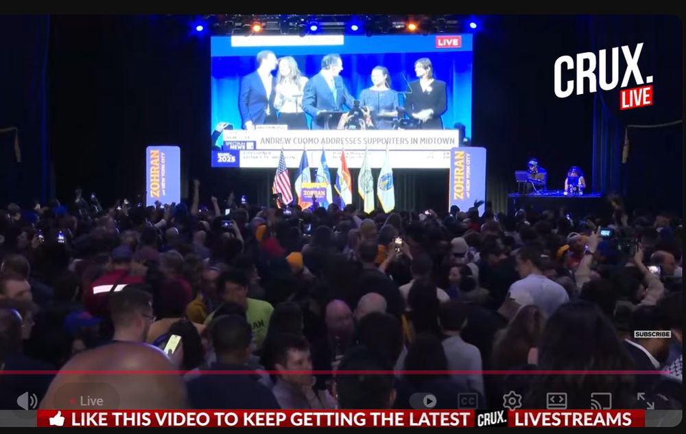Screenshot of the livestream from Mamdani HQ: the giant screen is showing Cuomo "addressing supporters in midtown" while people wait for the Mayor-Elect to speak