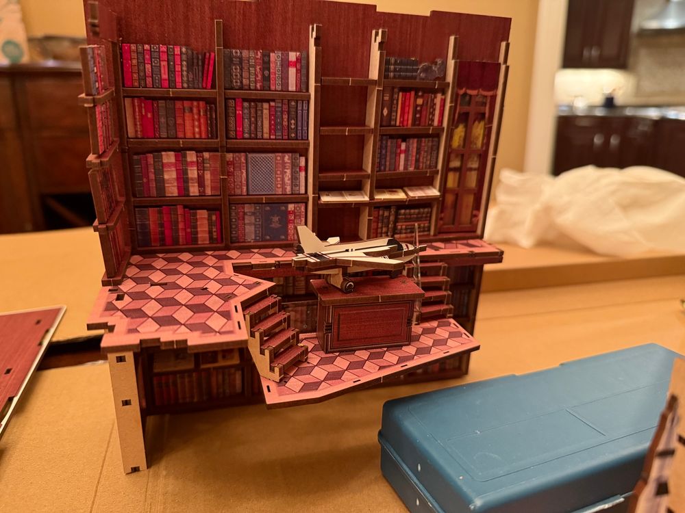Picture of a booknook assembly in progress showing the side panel with a wall of bookcases, a curtained window, and a model jet.