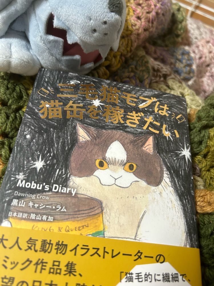 Dawning Crow作「摸布想自己賺罐罐 Mob's Diary(邦題/三毛猫モブは猫缶を稼ぎたい」の表紙を撮影した写真。猫缶を抱いた力強い顔つきの三毛猫のイラスト