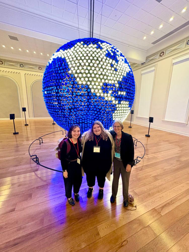 From left to right: Grace deMeurisse, Hannah Treadway, and Edith Kaan standing in front of a large illuminated blue and white globe at Planet Word in DC.