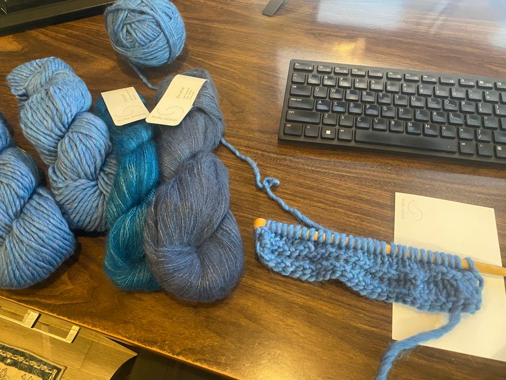 Assessing the 1 1/2” knitted blue scarf. Skeins of blue yarn lay on the desk next to project on left side. 