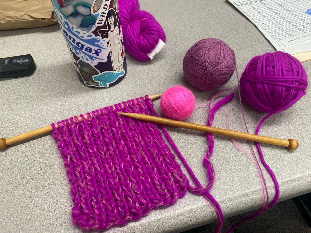 Three balls of yarn from bright pink to fuchsia, to mottled purple/pink sit on a desk with a scarf that’s about 5 inches long. 