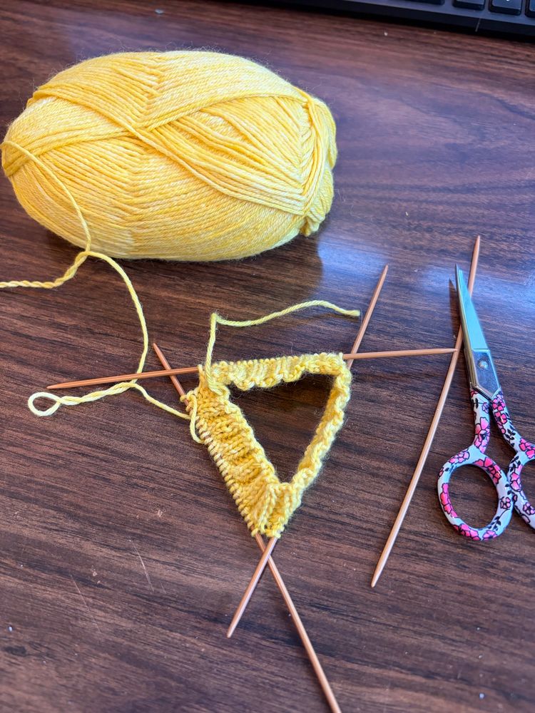 My first attempt at making socks. Three needles sit with a yellow 1/2” wide cuff and a pair of sewing scissors on a desk. 