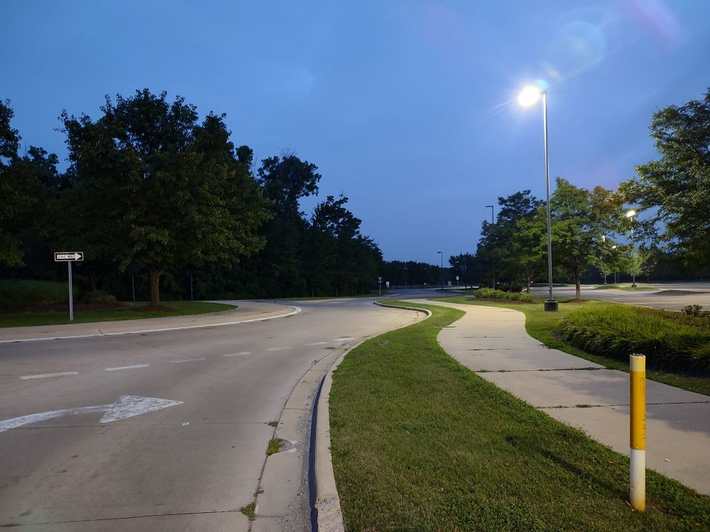 A road gently curves beneath a streetlamp, next to a sidewalk cutting through the grass. This stands under a cool blue evening sky.