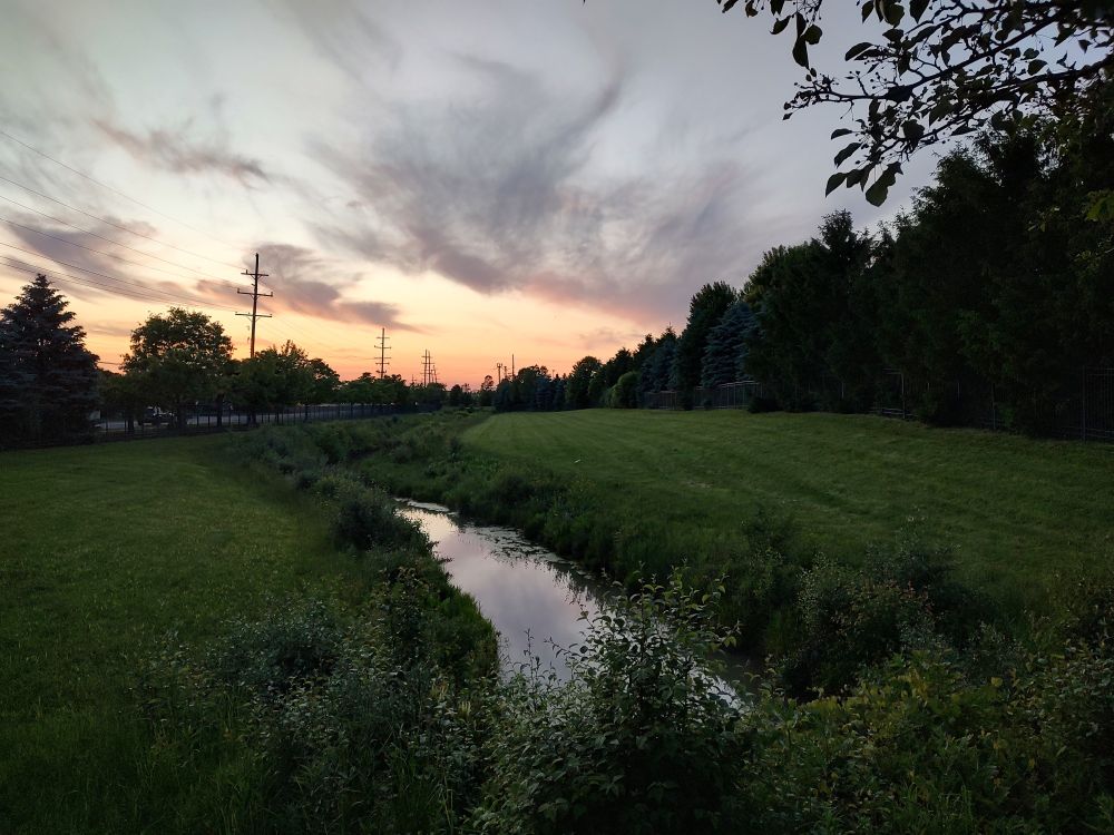 A stream under the same sunset and clouds, surrounded by deep green grass and trees