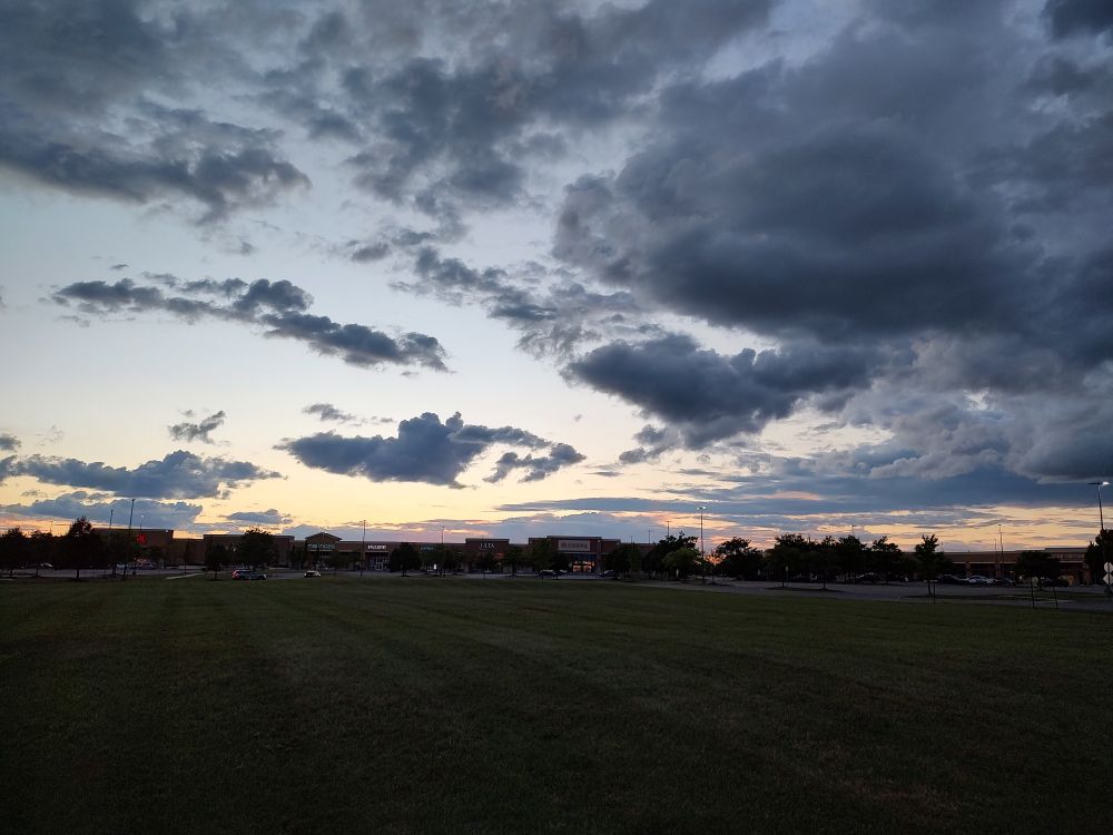 Dark grey clouds, in front of a blue sky, which quickly becomes a yellow sunset at the horizon. This rests over a small field near a shopping outlet.