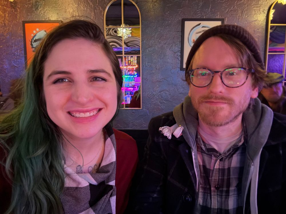 Smiling white woman with green hair and white bespectacled white man in beanie NOT smiling, colorful lights bar background 