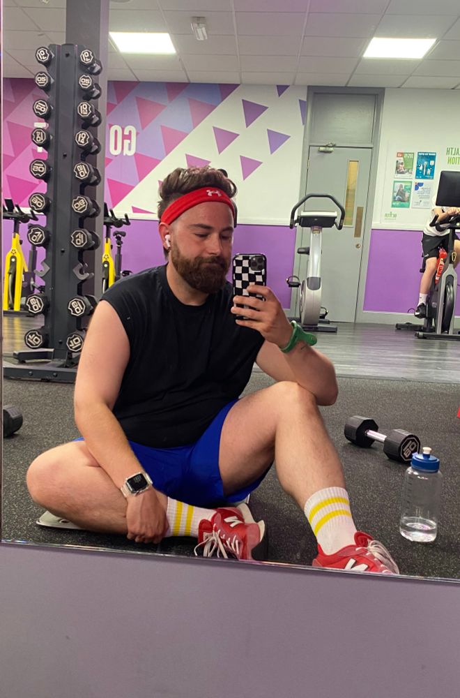 Bearded man sat on gym floor. Wearing black vest, blue shorts, red shoes and headband with striped yellow socks 