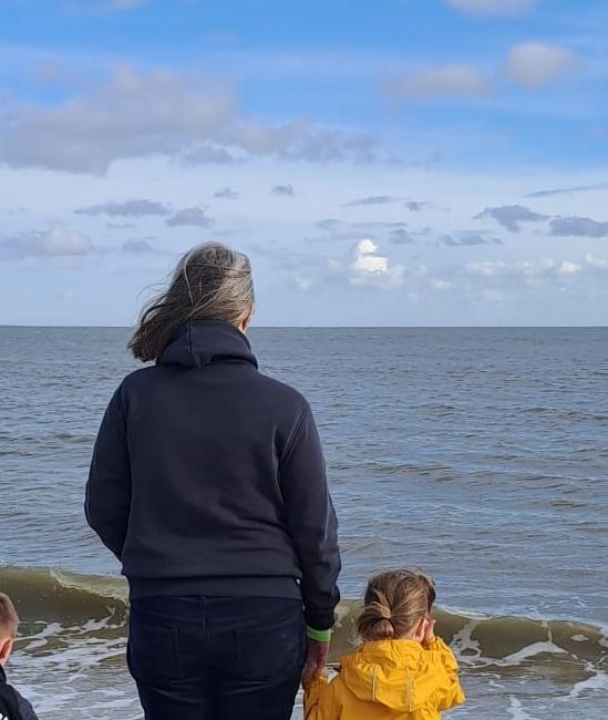 Blick auf die Nordsee. Direkt an der Wasserkante steht eine Frau in dunkelblauem Hoodie und dunkelblauer Hose. Ihre Haare wehen im Wind. An der Hand hält sie ein kleines Kind mit gelber Jacke. Beide blicken auf das Meer.
