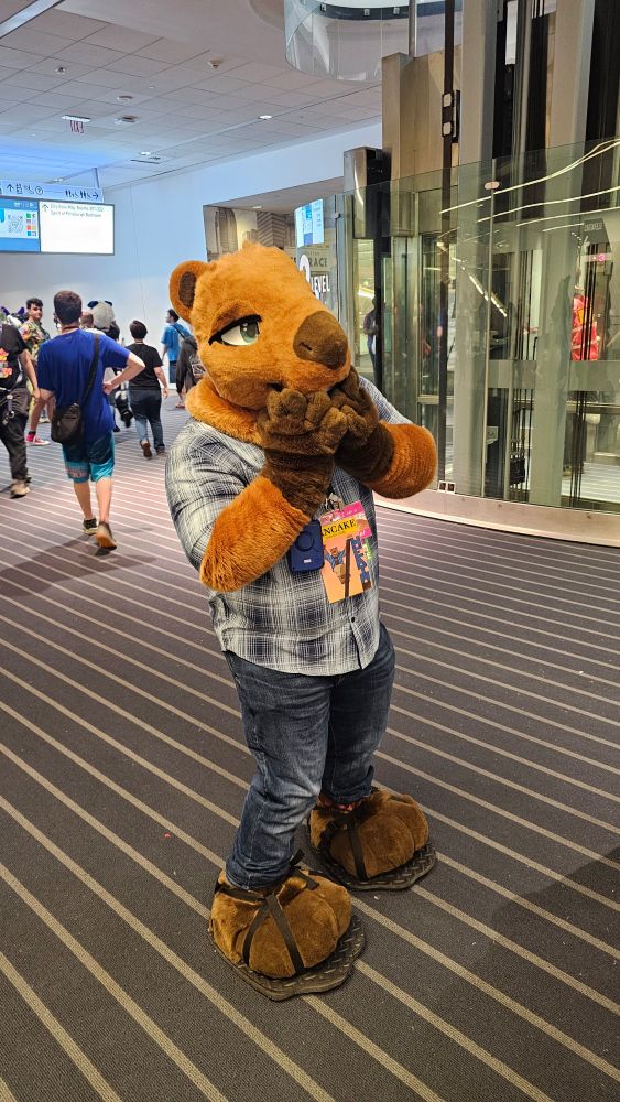 A capybara fursuiter on the convention floor. He covers his mouth with his hands 