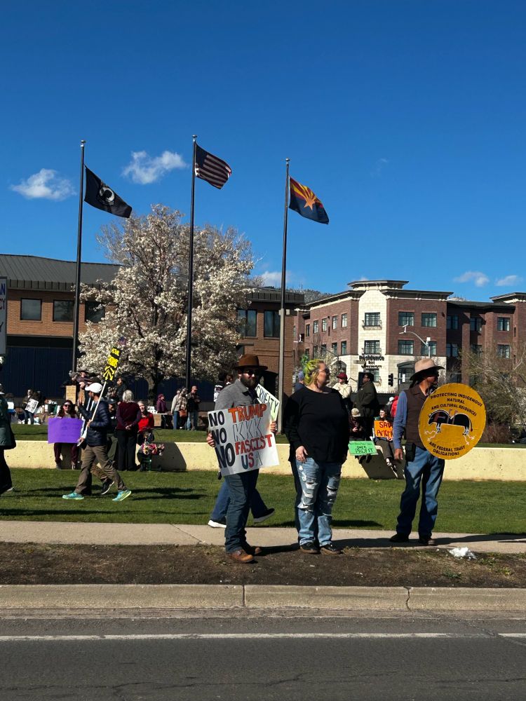 People stand with protest signs, including one about indigenous rights, in front of three flagpoles flying the US Flag, the Arizona flag, and a P.O.W. Flag.