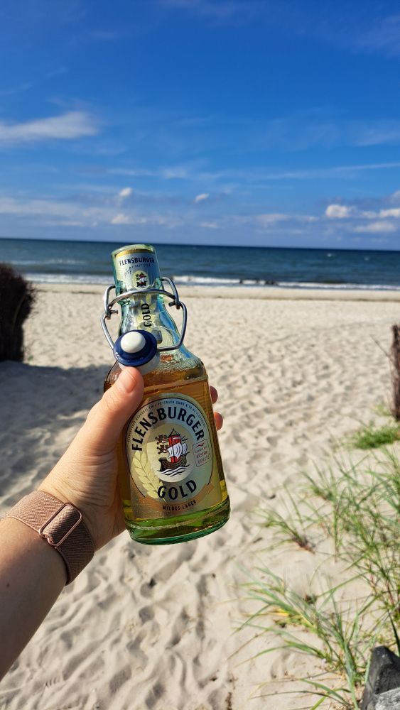 Eine Flasche Flensburger Gold in meiner linken Hand. Im Hintergrund Strand und das Meer 