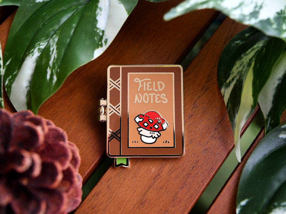 A hinged enamel pin that looks like a journal! It has a little mushroom on it, and above, it says "Field Notes"!