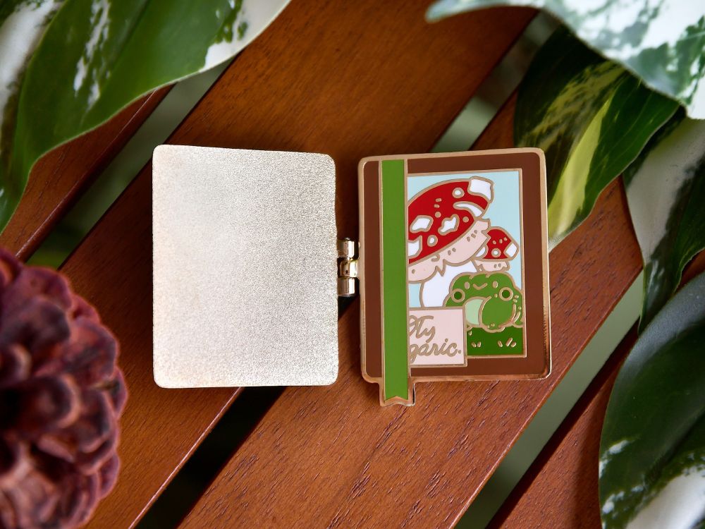 The same hinged enamel pin! It's open like a book, with Pond the frog, mushrooms, and a cute bookmark!