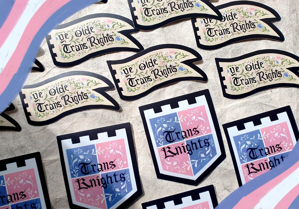 Photo of two sets of sticker designs. The first is a beige coloured banner decorated with medieval style flowers. It says "Ye Olde Trans Rights" in medieval inspired font. The second is a shield banner divided into pink and blue, separated by white. It says "Trans Knights" in a medieval style font.