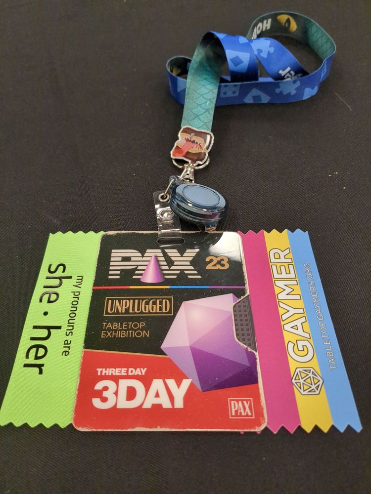A Pax unplugged badge is on a table with a green ribbon that reads my pronouns are: she/her on the left and a pansexual pride flag "gaymer" ribbon on the right.