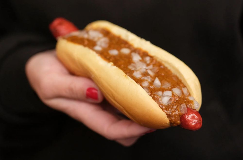 extremely haunting image of a woman's hand emerging from a black background, holding an unusually red hot dog smothered in the most watery chili you've ever seen