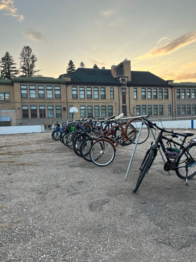 About 15-ish bikes hanging by their seats on a triangular bike rack, enclosed by a basketball court fence. There’s an old school in the background. 