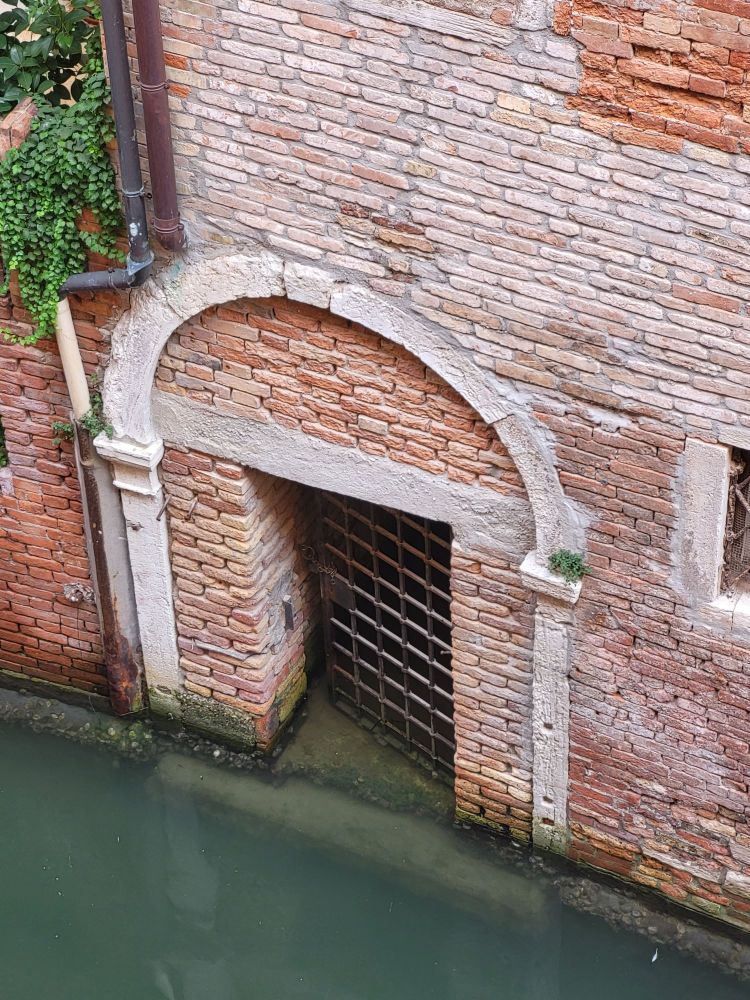 Looking down from a window in Venice at the a canal and the doorway in the wall opposite. The red brick wall has an iron gate at canal level that is partially submerged in water. The gate is ajar. The water is still and green, and a white stone step can be seen below the surface.