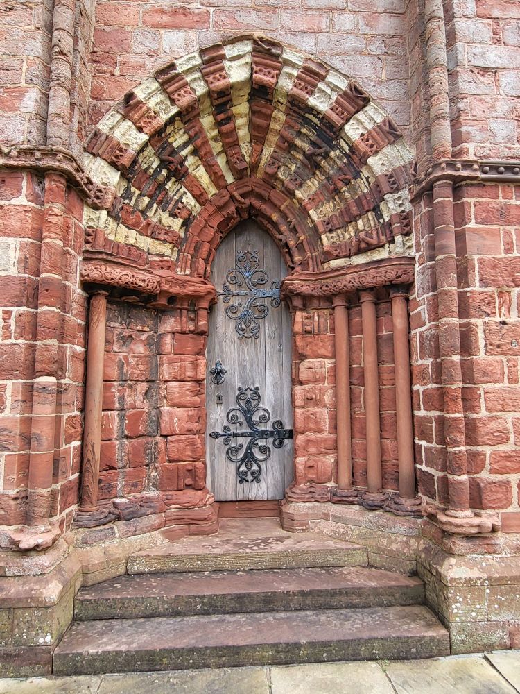A grey wooden door with elaborate, curling iron work straps, set into weathered red stone archway. The top of the arch has alternating blocks of red and white stone. Once ornately carved, the stone is heavily weathered and smooth. 