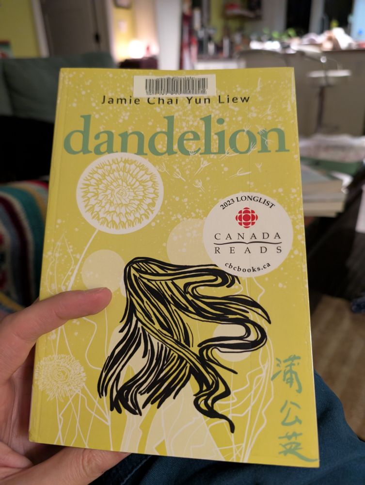 A hand holds the the cover of the book Dandelion by Jamie Chai Yun Liew. The cover is light green with the title across the top, a woman's long dark hair is drawn in, swirling. There is a Canada Reads 2024 Longlist sticker on the cover.