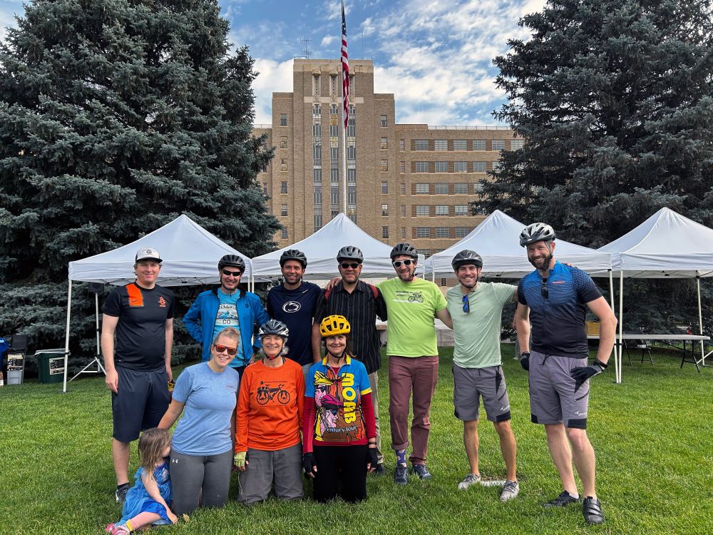 A group of seven men, three women, and a child, smiling with their arms around each other. They're wearing bike helmets after completing Bike to Work Day.