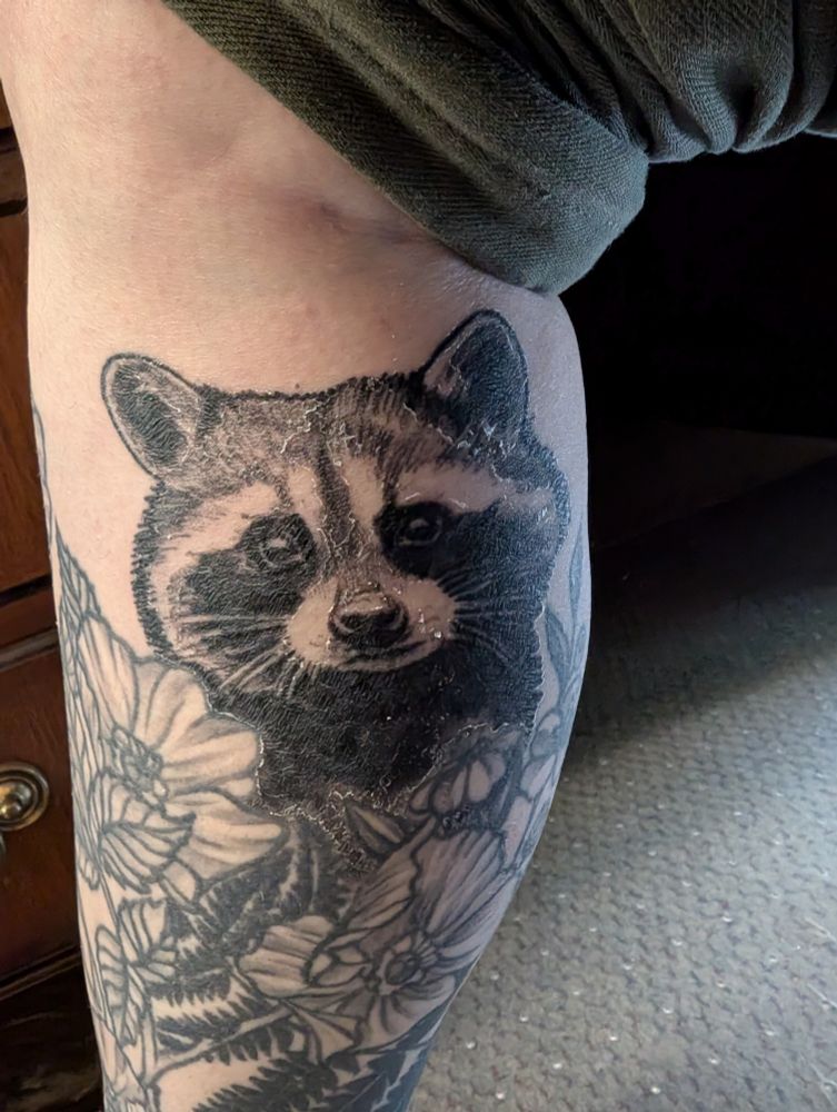 Tattoo of a raccoon on a calf surrounded by flowers
