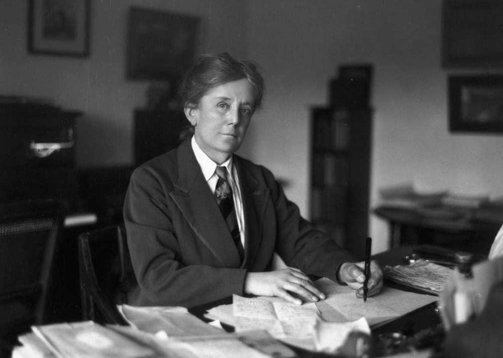 Black and white portrait of Ethel Smyth from British Music Collection. Ethel wears formal suit, white shirt and a tie, sitting at table that is covered with papers. 