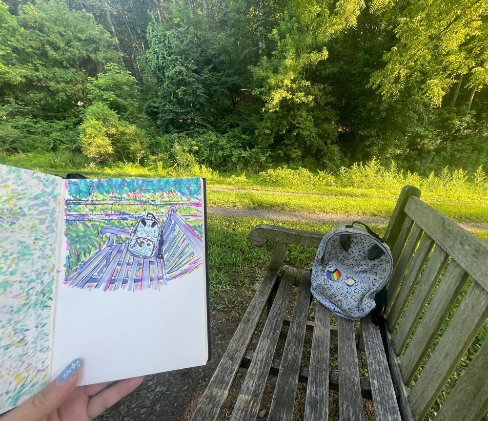 We see a marker sketch of a scene held up to it. In both, as a viewer we are sitting on a bench viewing a back pack in front of a path through grass. The backpack has some pride pins and is light blue with a cartoon cat pattern and two “ears” on top of the bag. The marker sketch is colorful with bright, random colors. 