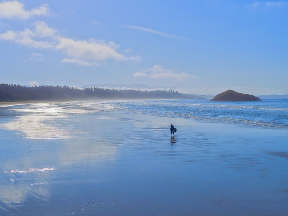clouds and blue sky reflected on the wet endless sands with a sole surfer walking almond the beach at low tide. 