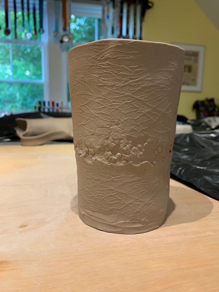 A wet grey clay pint glass with raised texture resembling fine twigs or grass with a band of rougher, random circular texture running horizontally around the center