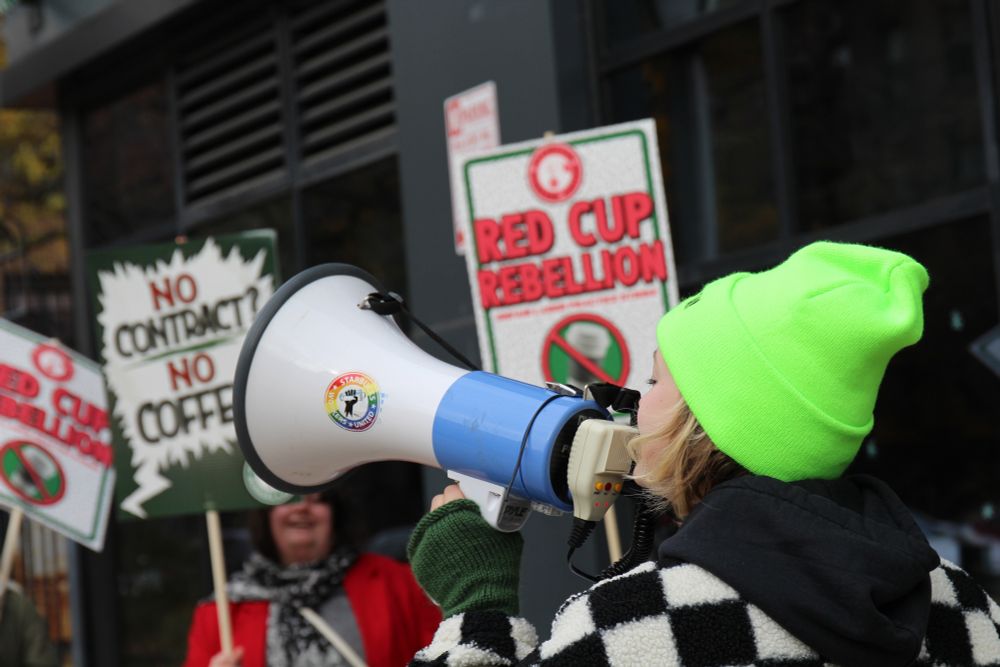 A strike captain rallying Starbucks workers in front of a store holding signage that reads "No Contract? No Coffee," and "Red Cup Rebellion."