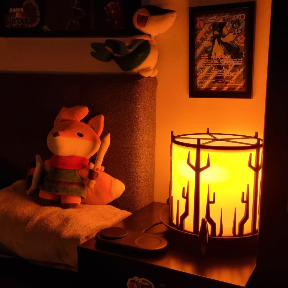 A picture of a bedside table, with one of the Outer Wilds-themed lanterns on top, lit up in a cozy, warm light.