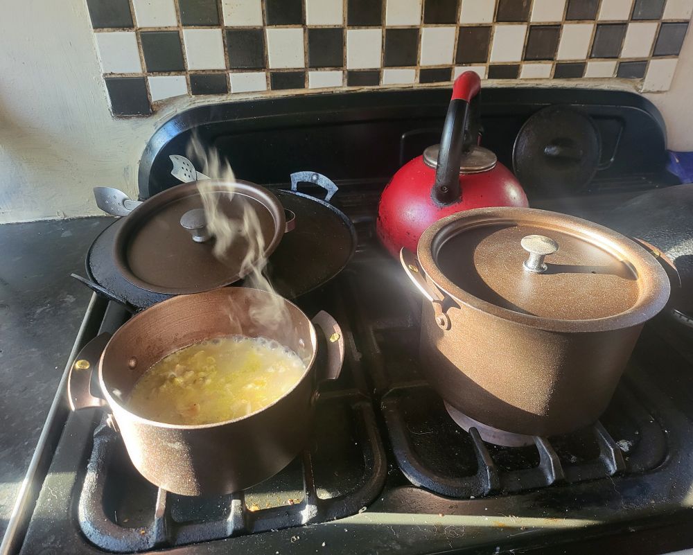 Two brand new iron stew pans sit on the front row of a black gas hob. The leftmost one contains leek and potato soup from which a whisp of steam ascends. Behind the stew pans are a well-used iron prospector pan and a red kettle. Behind them are black and white chequered tiles.