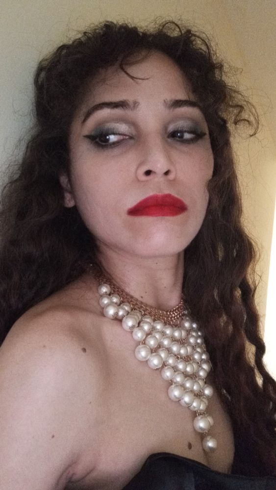 Uma mulher de cabelos cacheados com uma parte presa para trás, batom vermelho, está maquiada, com uma expressão séria e um colar com um cordão dourado que sustenta fileiras de pérolas na posição vertical. A mulher olha para o lado, assim como está posicionada e suas costas estão grudadas na parede