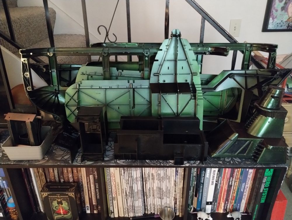 A disassembled toy cruise ship freshly spray painted & sitting on bookshelf of role playing game books waiting for the next step in its refurbishing. 