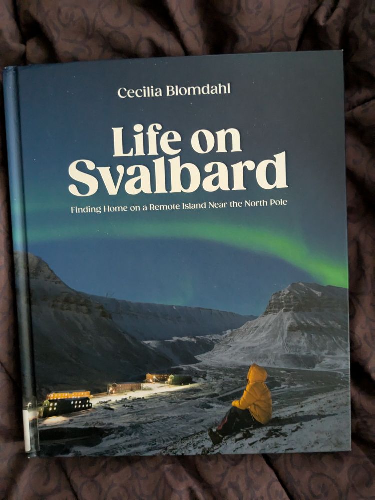 Cover of the book Life on Svalbard Finding home on a remote island near the north pole. Shows a building, a mountain or glacier with lots of snow and a person wearing black pants and a yellow jacket with the hood up looking out at the scenery around them.