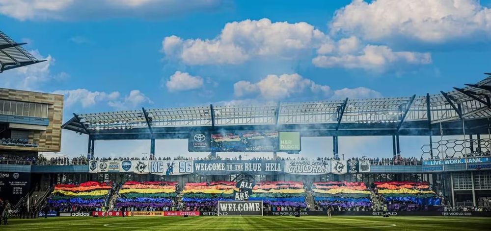 A photo of the "Cauldron" supporters section at Children's Mercy Park, home of Sporting Kansas City. Long banners span the crowd, left to right, forming a rainbow. At center, three signs state "All Are Welcome"

Photo provided by KC Cauldron