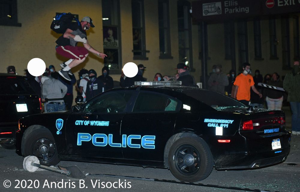 Cop car surrounded by sparse crowd, while a person with shorts and a backpack is midair above the hood, prepared to stomp down with both feet.