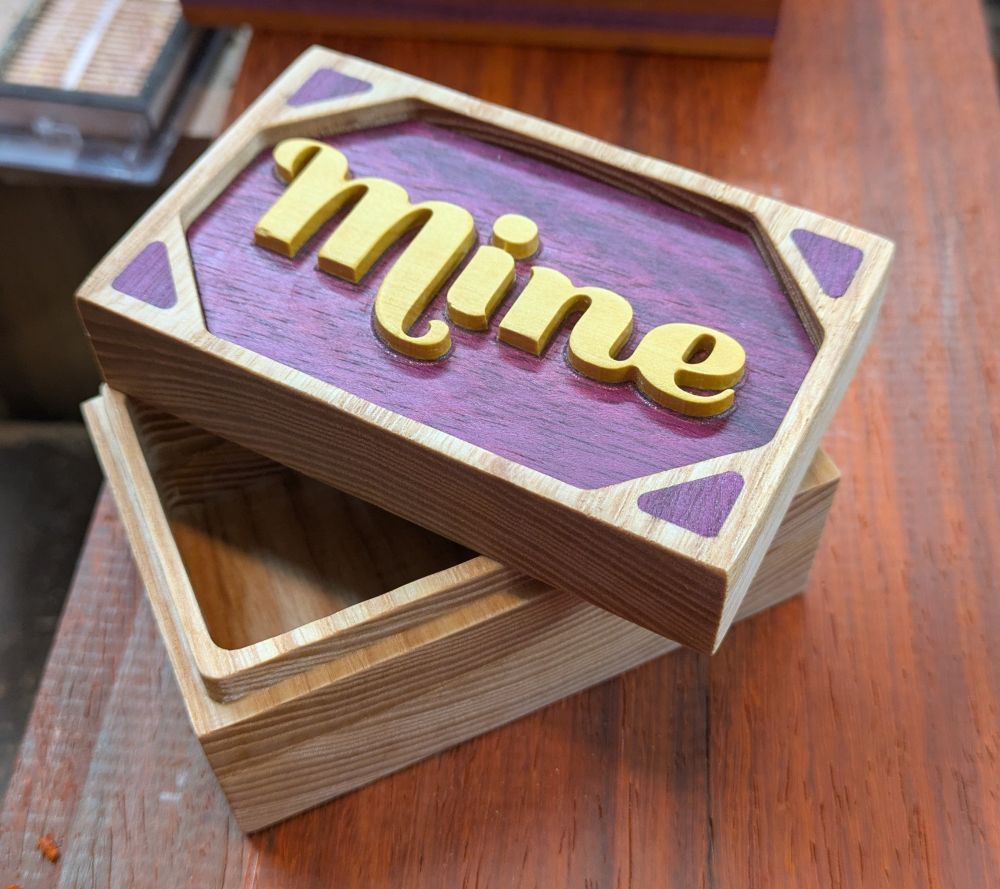 A small wooden box I light wood with purple inlay and the word "Mine" carved into the lid in a yellow wood