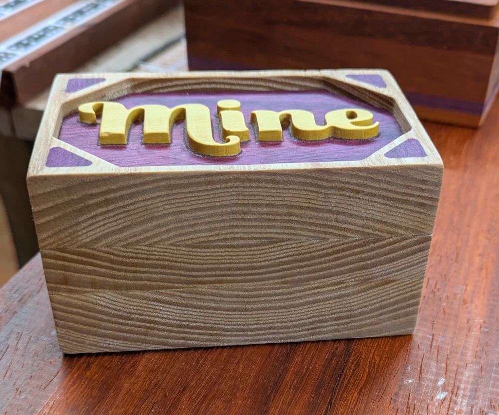 A small wooden box I light wood with purple inlay and the word "Mine" carved into the lid in a yellow wood
