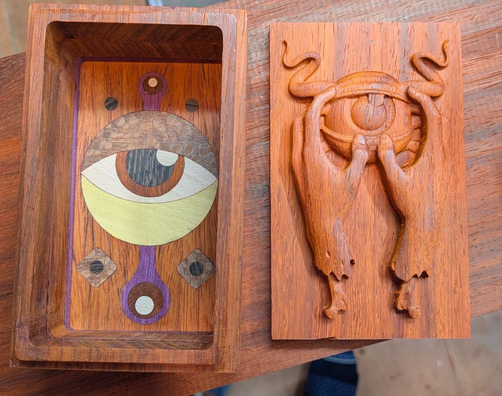A wooden box with inlay of eye in different colored wood at the bottom - it's lid sits next to it with a 3d carving of two severed hands holding an eye with horns carved into the wood lid