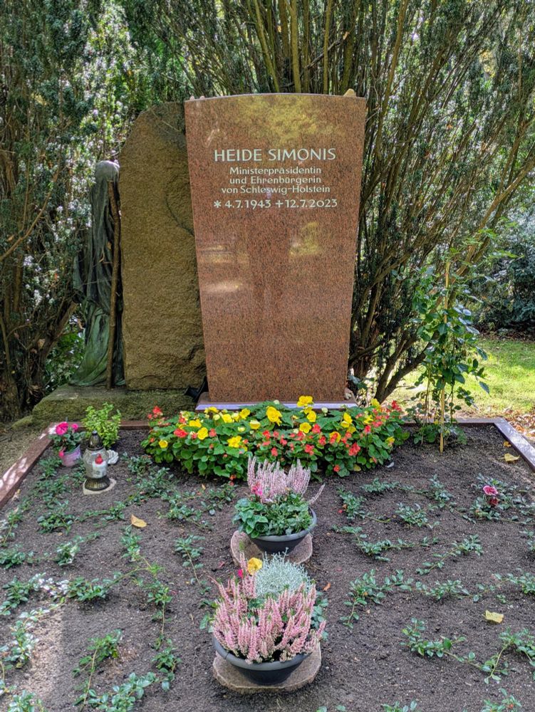 Einfacher brauner Grabstein mit Inschrift, davor ein paar Blumen und Pflanzschalen