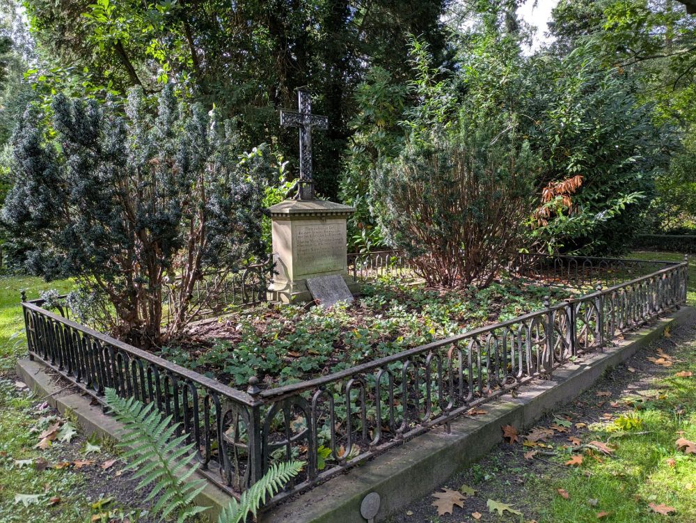 Grabstätte, von niedrigem Metallzaun umgeben. Zentral ein Metallkreuz auf einem Steinsockel. Zwei Büsche flankieren das Kreuz.