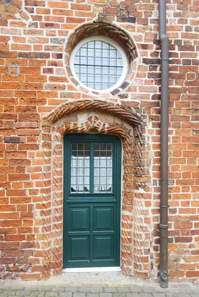 Eine dunkelgrüne Tür in einer Backsteinmauer. Darüber ein rundes Fenster.