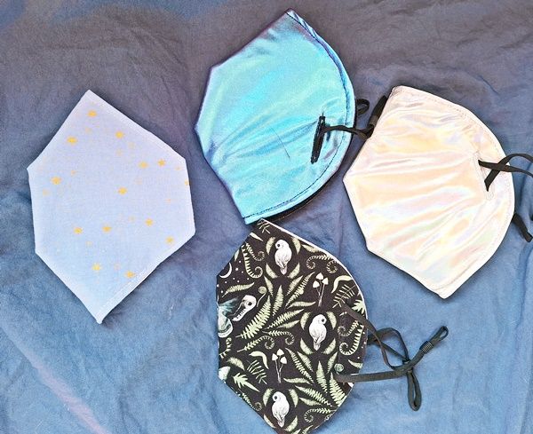 A set of four N95 masks, each covered in a fabric, one periwinkle with gold stars, one shiny blue, one shiny silver, and one black with owls, ferns and moths. 