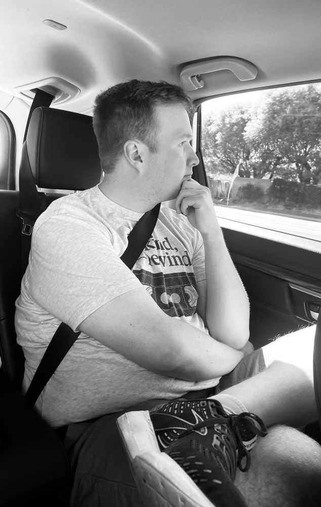 A black-and-white photograph of a young man in the backseat of a car, looking at the window, pondering at the view.