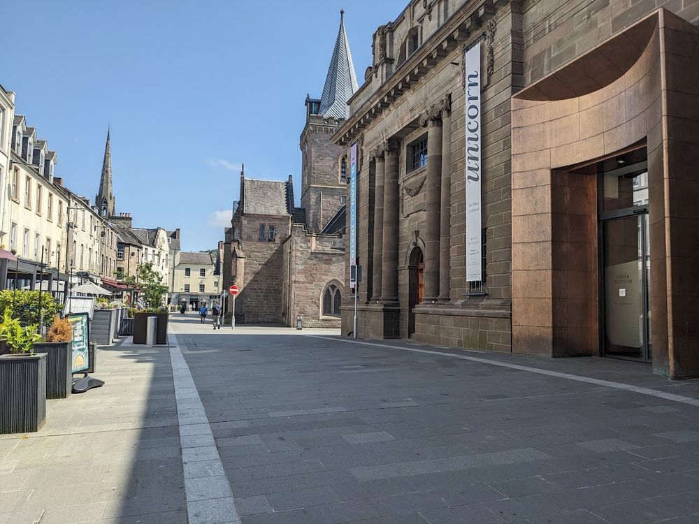 Sun shining on the stone buildings of Perth. A paved pedestrianised area with cafes on the left and the museum on the right and the St Johns Kirk with its spire behind.