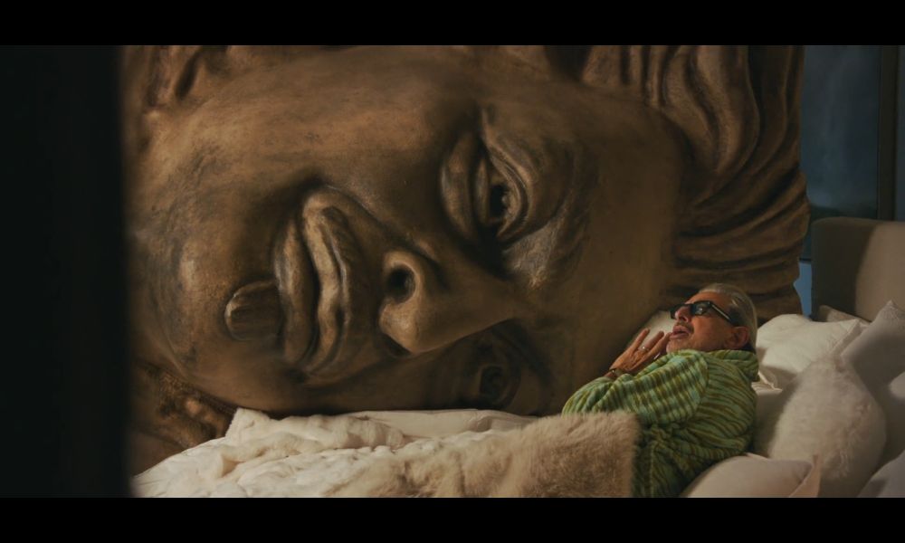 Jeff goldbloom laying in bed with a massive stone statue of his face from the set of wicked