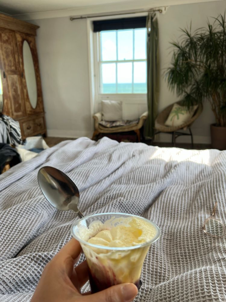 A view of a bright bedroom taken from bed. In the background is a window with a beautiful sea view. In the foreground is a hand holding a small dessert trifle and spoon. 